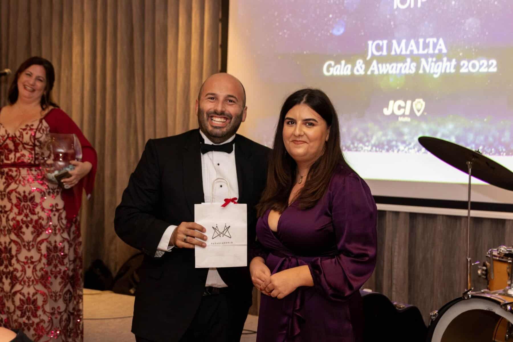 320245466_947490599562045_2980159045296685064_n Award-winning man in tuxedo and woman in purple dress at a formal event with a large screen displaying "JCI Malta Gala & Awards Night 2022" in the background.