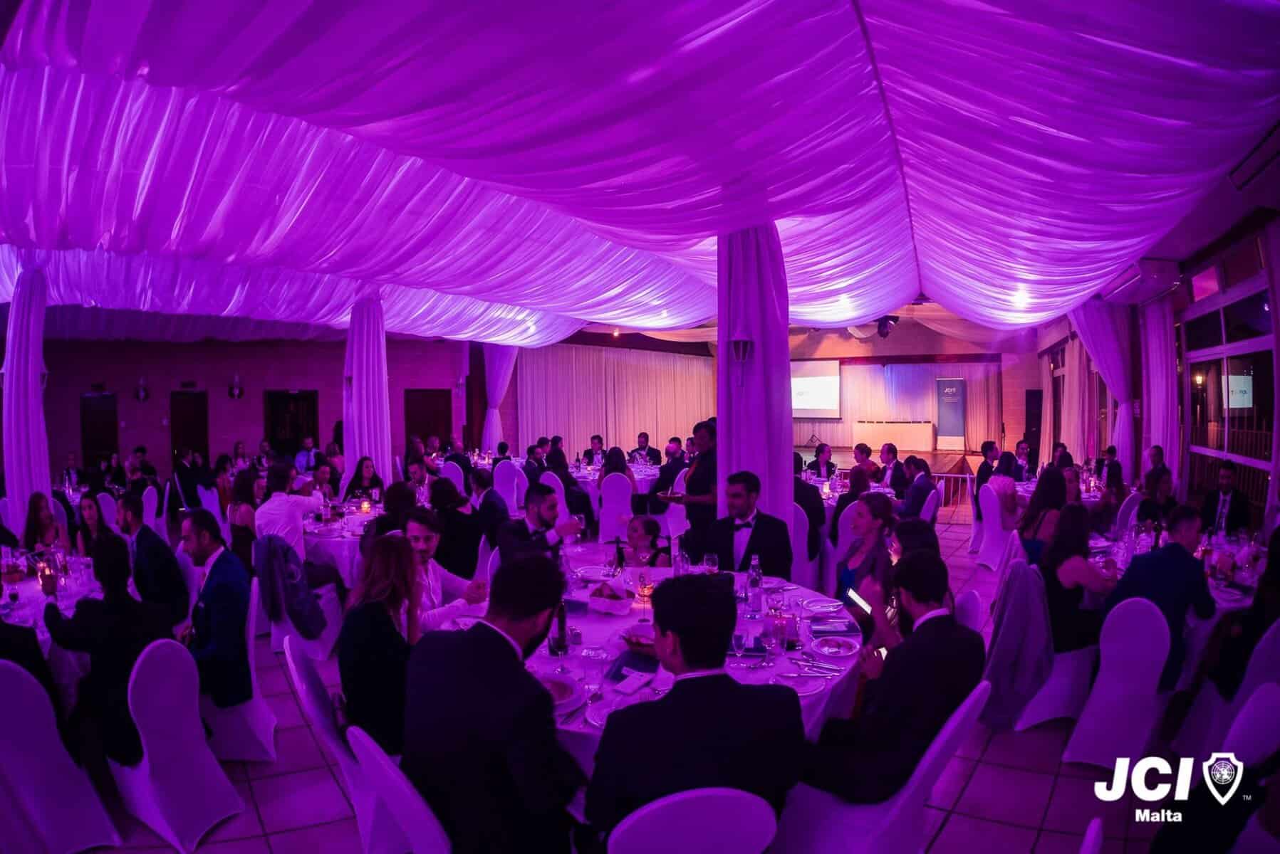 JCI Awards night Elegant banquet hall decorated with purple lighting, hosting formal event with seated guests in suits and dresses, creating a sophisticated and lively atmosphere for celebration or gathering.