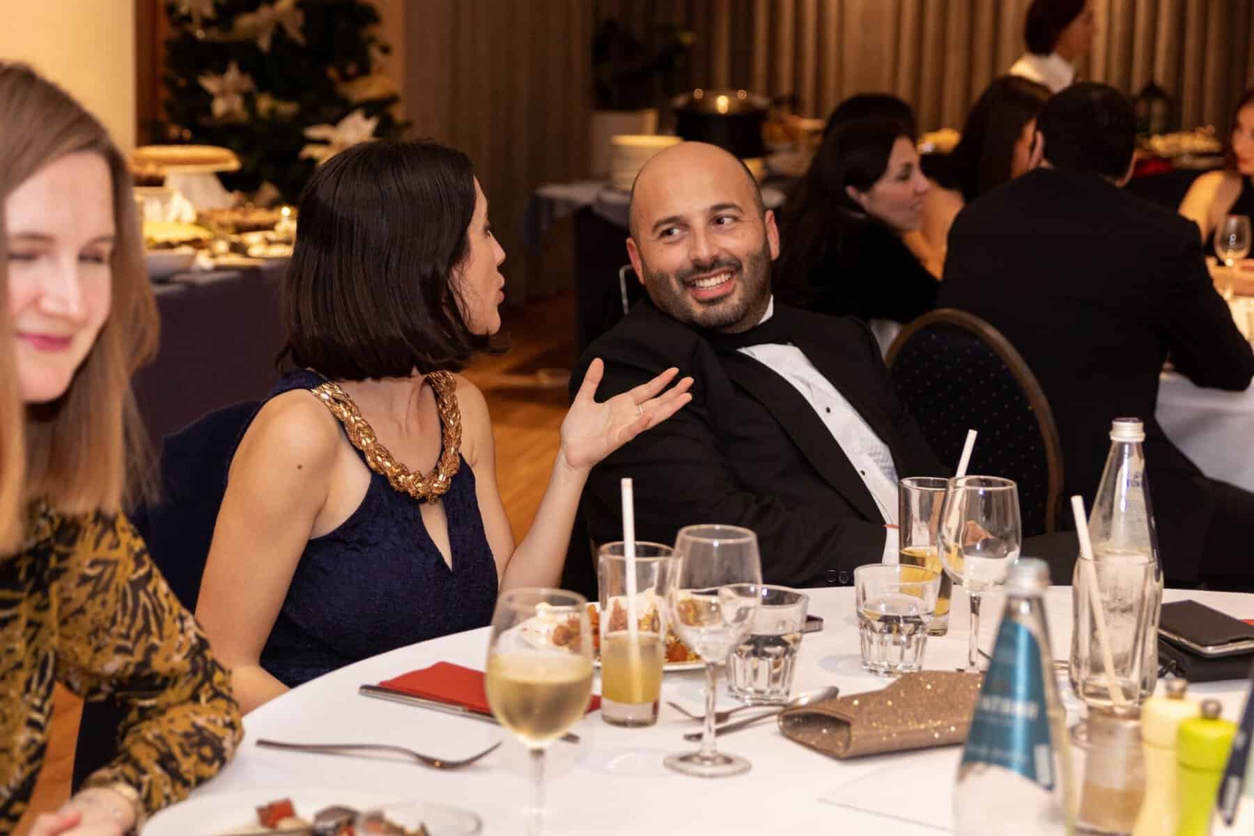 Young Entrepreneur Accomplishment nomination A man in a tuxedo smiling and engaging in conversation with a woman wearing a navy blue dress and gold chain necklace at a formal dinner event. Other guests are seated at the table, enjoying drinks and food.