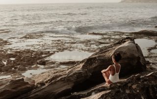 Reflective woman meditating on rocks by the ocean, practicing mindfulness and self-awareness for mental clarity and emotional well-being.