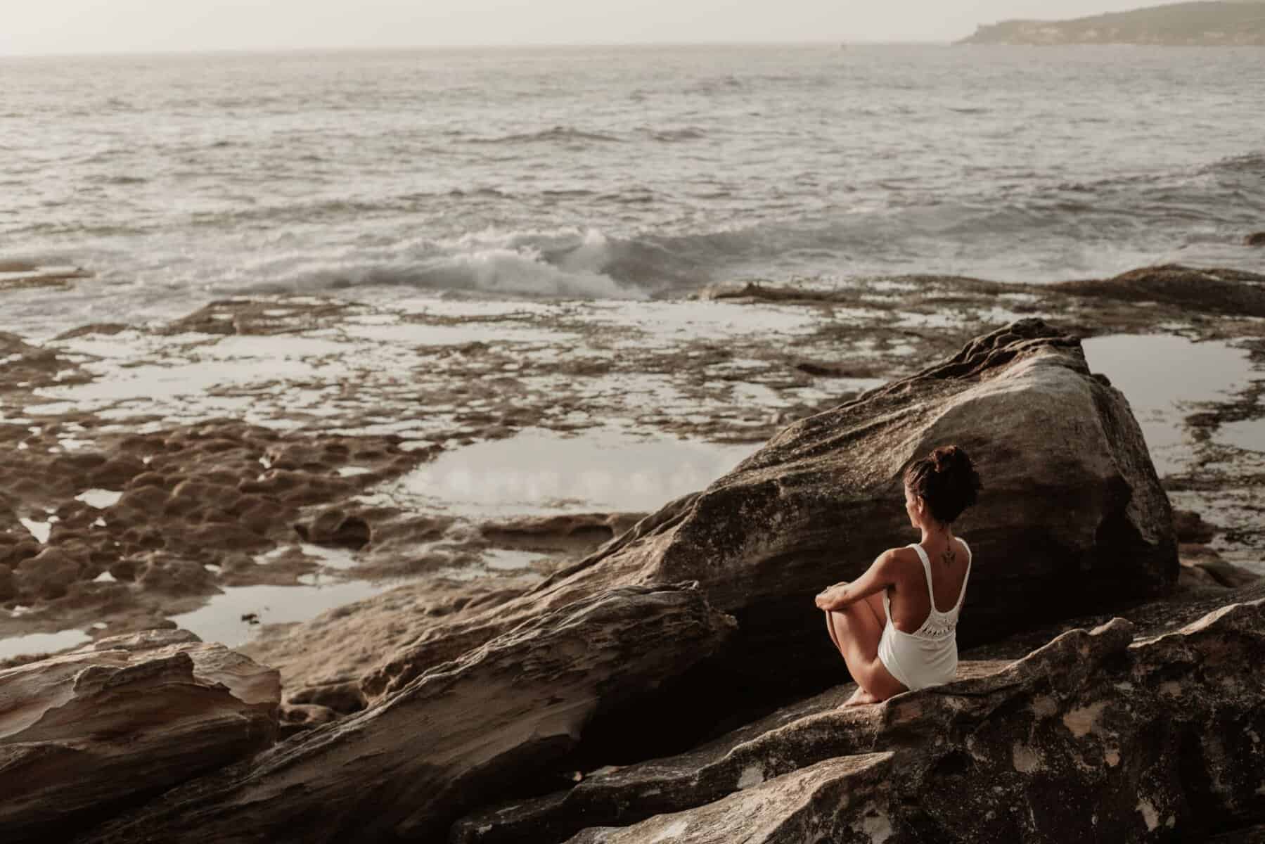 Reflective woman meditating on rocks by the ocean, practicing mindfulness and self-awareness for mental clarity and emotional well-being.