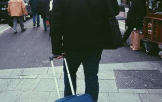 A person standing at a busy city street corner, pulling a rolling suitcase, dressed in dark clothing, with other pedestrians and street vendors around.