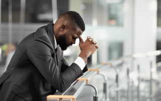 Deeply distressed man in a business suit holding his head in frustration while leaning on a railing in a modern office lobby or public space, experiencing emotional stress or anxiety.