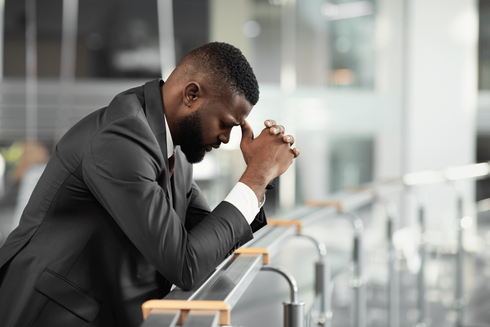 A manager in an office environment. He is leaning his elbows on a glass railing with his hands to his face. He is showing facial features of worry.