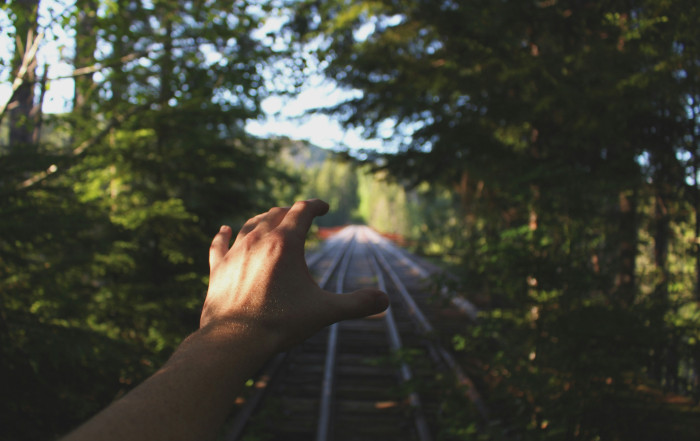 It showcases a hand reaching out in the air. The scenery behind the hand is a train track, surrounded by trees on the sides.