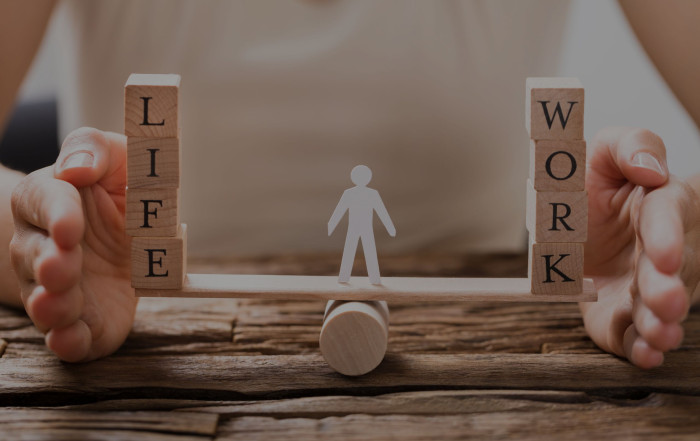 a white figure in the middle of a see saw, one side there is the word life and the other side there is the word work representing a balanced life.