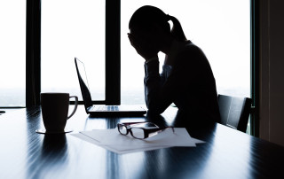 Overwhelmed woman holding her head while sitting at a desk with a laptop, glasses, and papers, indicating stress or mental health struggles in a professional or personal context.