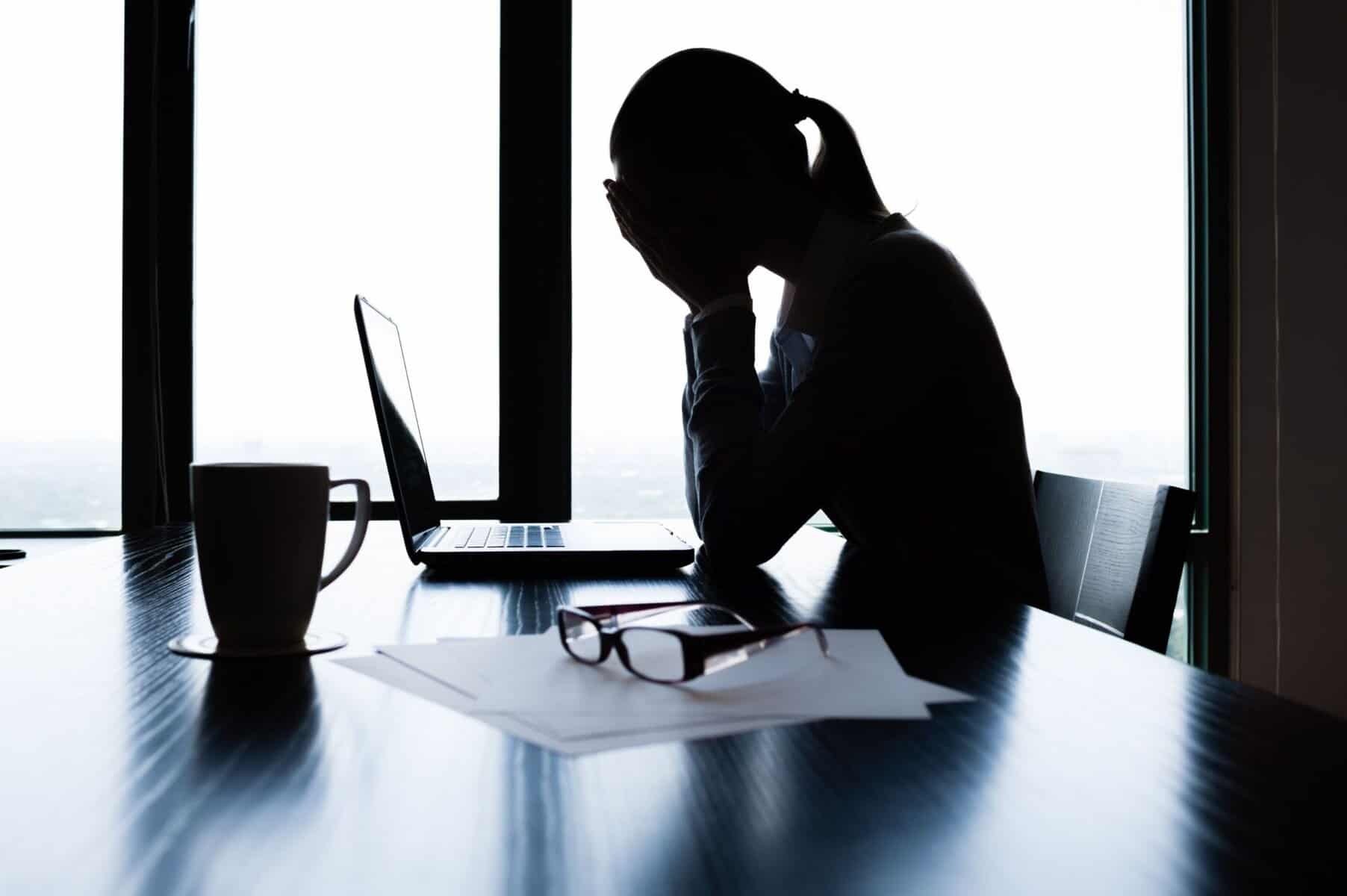 Overwhelmed woman holding her head while sitting at a desk with a laptop, glasses, and papers, indicating stress or mental health struggles in a professional or personal context.