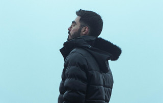 Dark-haired man in a black winter jacket with fur-lined hood standing outdoors against a cloudy sky, representing mindfulness and stress relief, emphasizing calmness and mental clarity in challenging times.