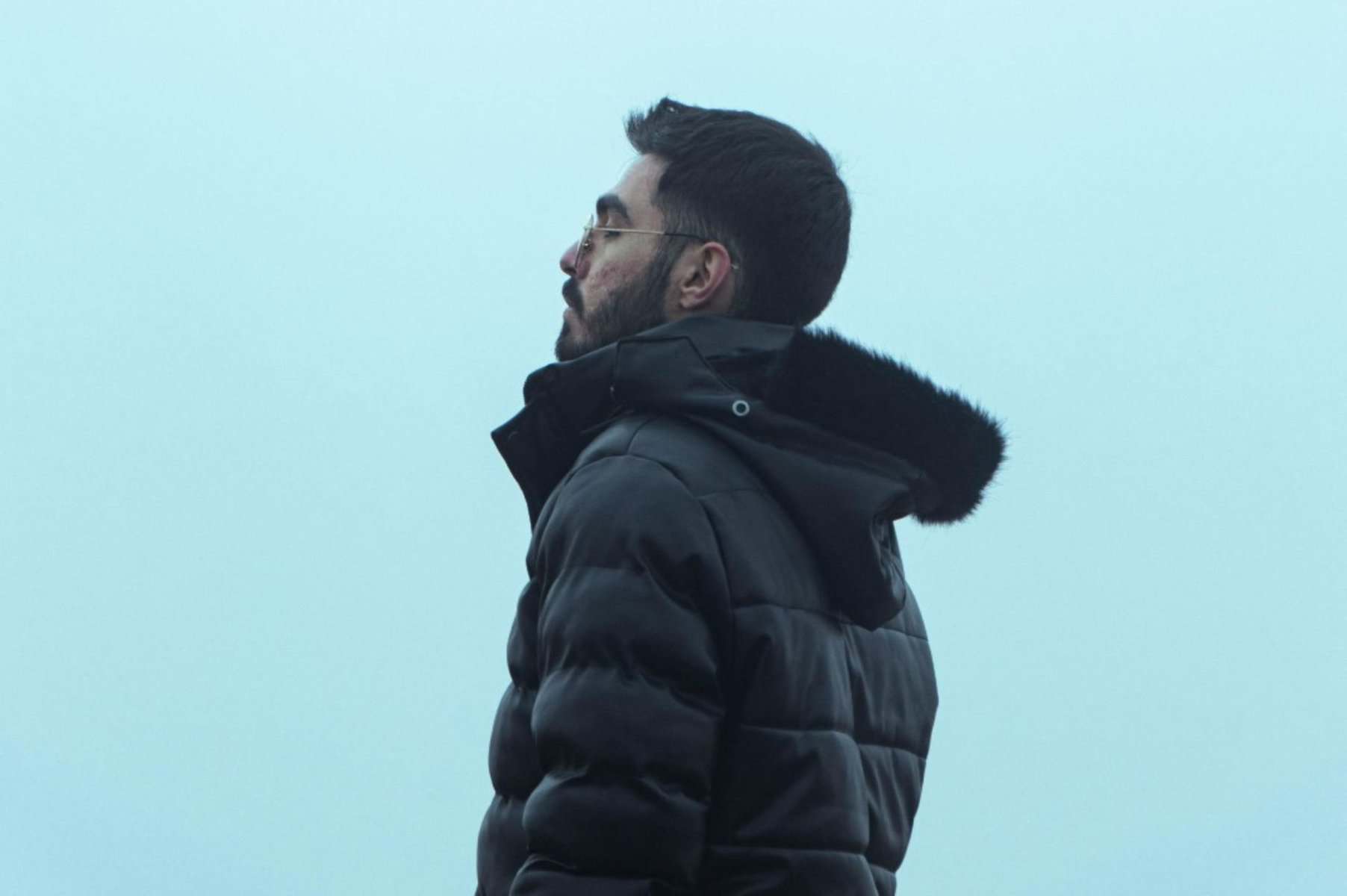 Dark-haired man in a black winter jacket with fur-lined hood standing outdoors against a cloudy sky, representing mindfulness and stress relief, emphasizing calmness and mental clarity in challenging times.
